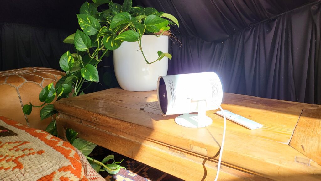 White Samsung portable projector sitting on a wooden table with a very green ivy plant next to it.