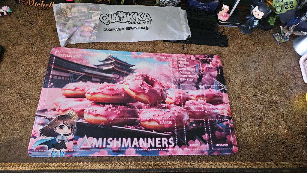 Magic: The Gathering playmat with custom MishManners branding. The Quokka Mousepads packaging is sitting just above the playmat.