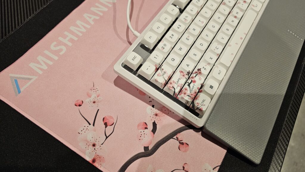 Minimalist mousemat light pink with cherry blossoms in the corner and a MishManners logo in the top left hand corner. The mousemat is sitting on top of another black mousepad.