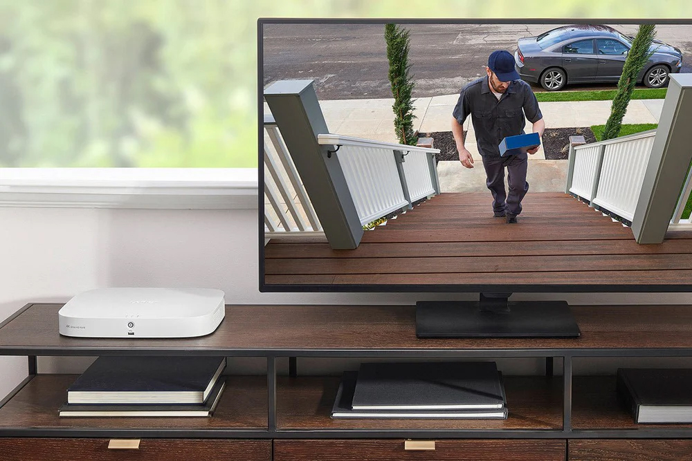 White Lorex security device sitting on a wooden table next to a monitor that's showing a high definition image of a person delivering a package to the front door.