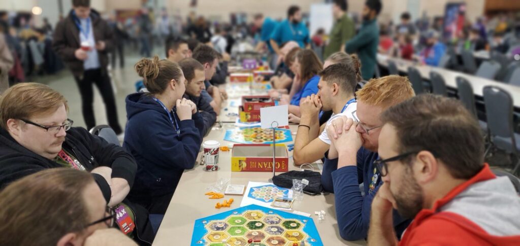 一排桌子上摆放着多张《卡坦岛》棋盘，玩家正在互相竞争。