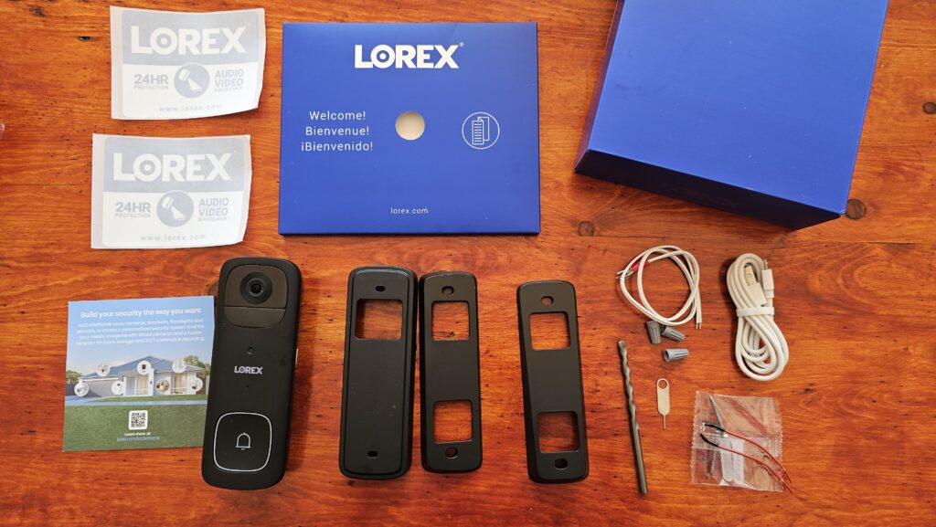 Lorex 2K Battery Video Doorbell out of the box showing doorbell, mounting bracket, vertical and horizontal inserts, drill bit, hard wired wiring, USB cable, stickers, and instruction manual