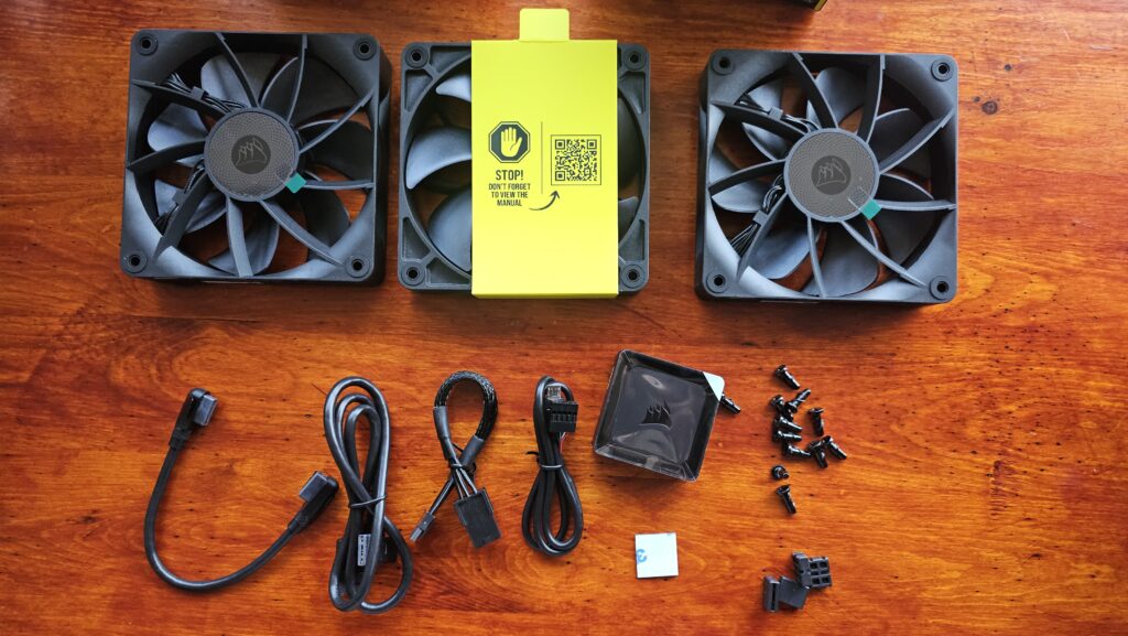 Three black fans, iCUE controller, USB and power connectors, two cable options for connecting the fans, screws, and inter-fan connectors. All black.