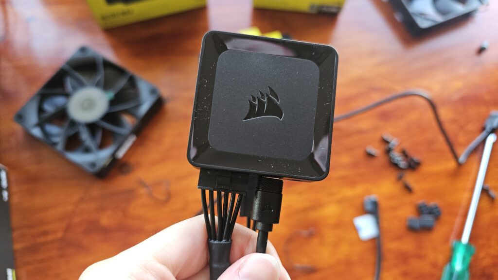 Square, black, iCUE hub with the Corsair logo in the middle. There are two cables coming out of it.